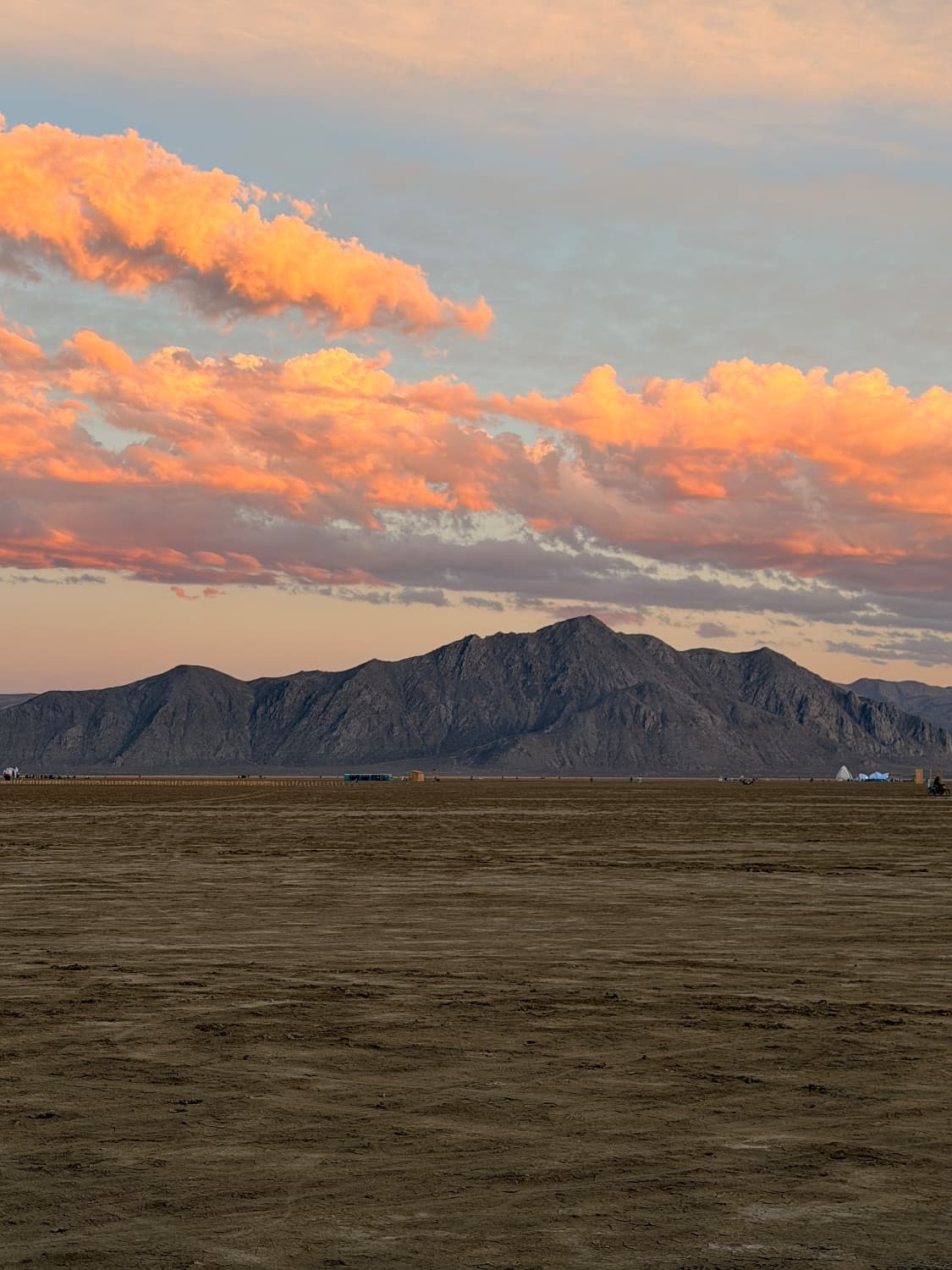 Sunrise over the Black Rock Desert, pink and orange clouds lit above the mountain range, with open playa stretching to the horizon and a few distant camps along it