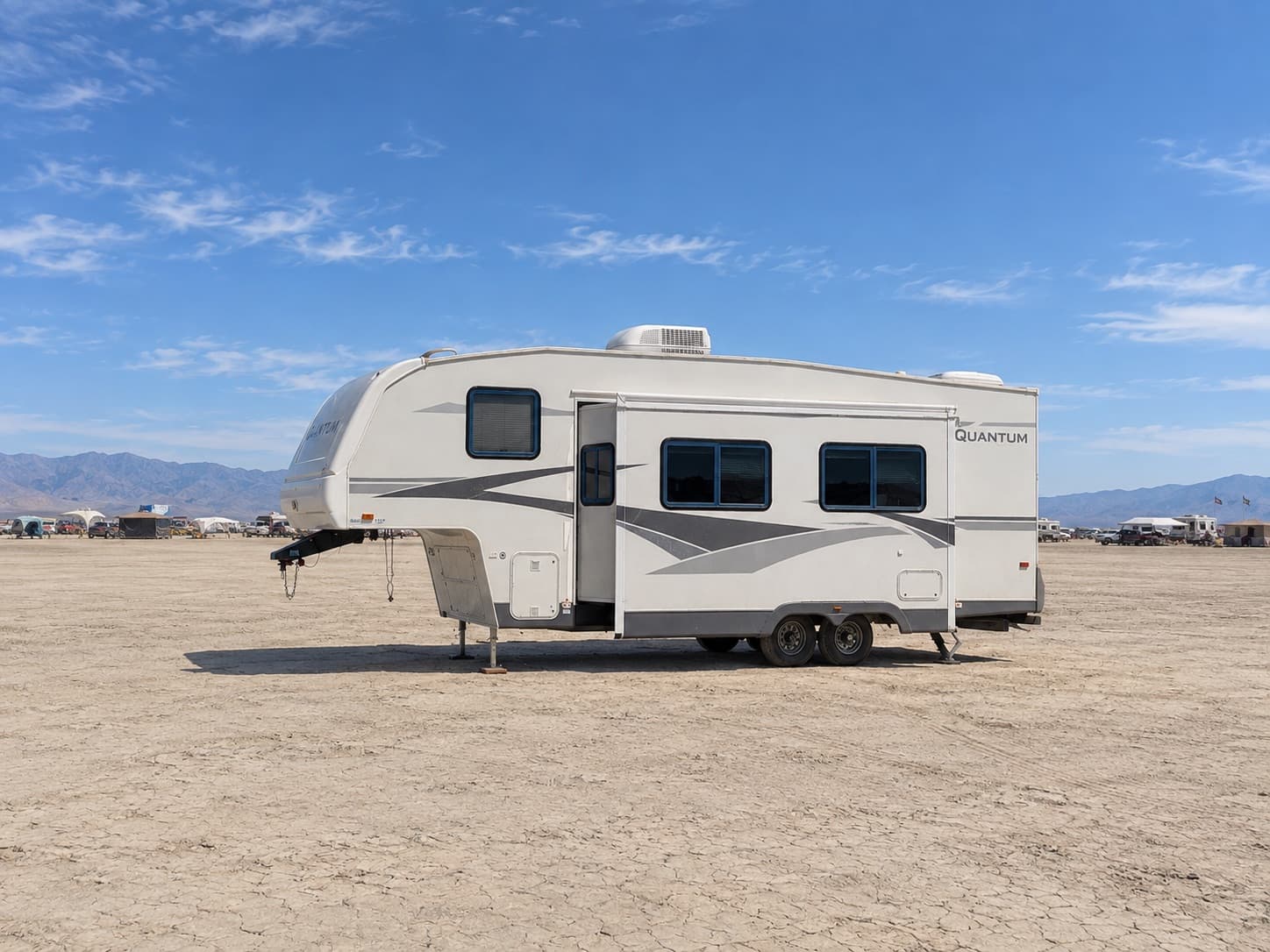 The Quantum, a white 25-foot Fleetwood Terry Quantum fifth wheel, levelled on the open playa with its slide-out extended and its roof air conditioner on top, mountains and distant camps behind it